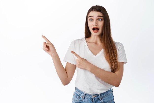 Excited and surprised young woman gasping wondered pointing and looking aside left at copy space shouting from amazement and joy standing in tshirt against white background