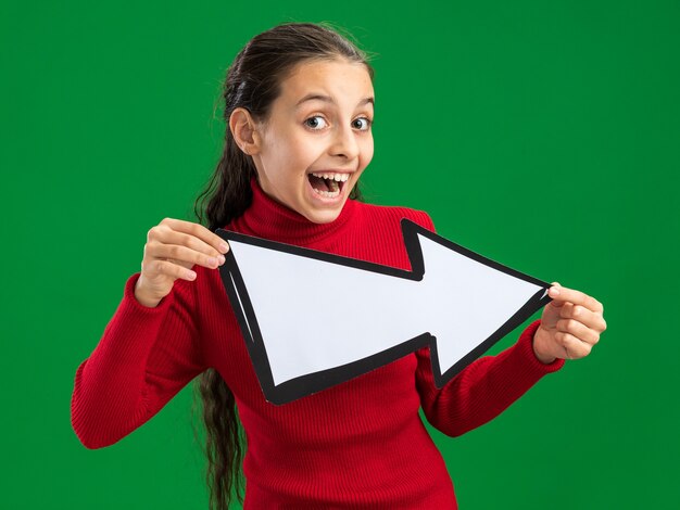 Excited teenage girl holding arrow mark pointing at side looking at camera isolated on green wall