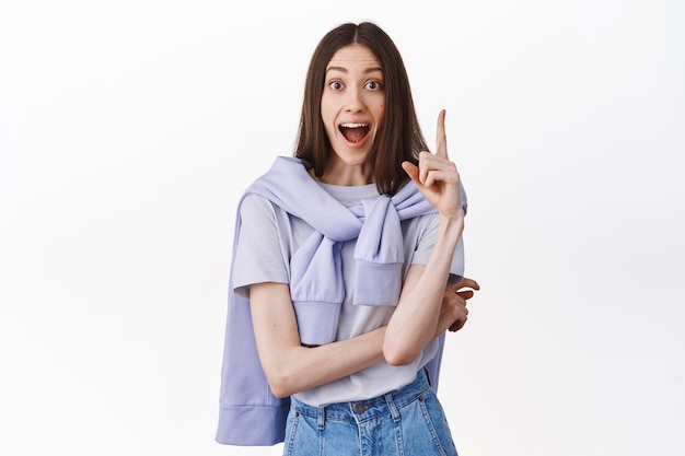 Excited, thrilled brunette girl screams while raising finger in eureka sign, have great plan, think up solution, saying her suggestion, standing over white wall