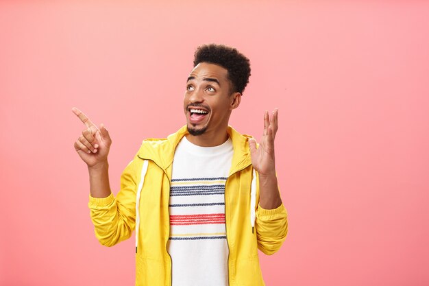 Excited and thrilled dark-skinned young male with beard and afro hairstyle raising palm in amazement pose pointing and looking at upper left corner astonished and delighted over pink wall