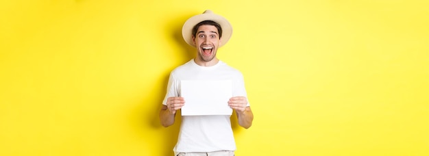 Free photo excited tourist showing your logo or sign on blank piece of paper smiling amazed standing against ye