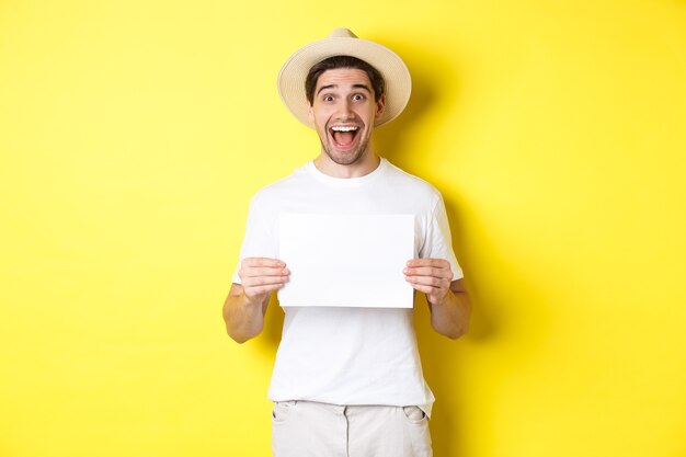 Excited tourist showing your logo or sign on blank piece of paper, smiling amazed, standing against yellow background.