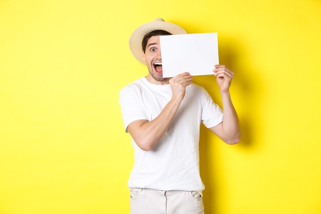 Excited tourist on vacation showing blank piece of paper for your logo, holding sign near face and smiling, standing against yellow background