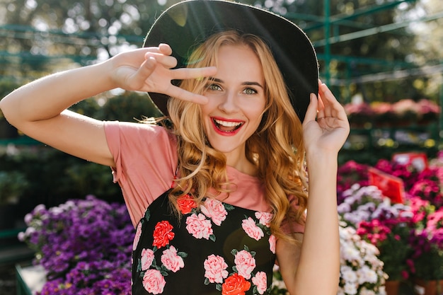 Excited white woman with blinde hair having fun in greenhouse. Portrait of joyful woman dancing in frint of flowers.