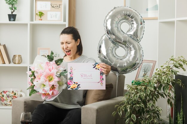 Excited with closed eyes beautiful woman on happy women day holding bouquet with postcard sitting on armchair in living room