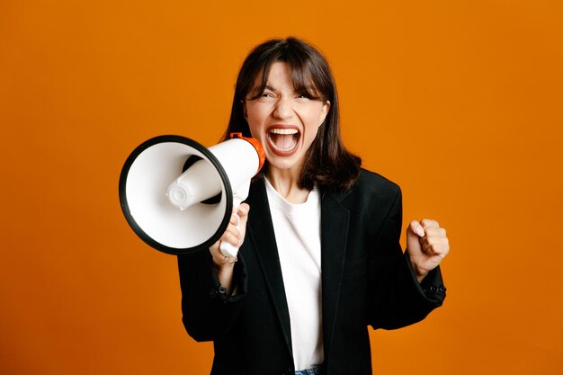 Excited with closed eyes speaks on loudspeaker young beautiful female wearing black jacket isolated on orange background