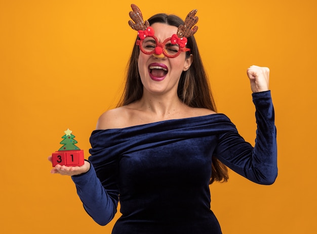 Взволнованная, с закрытыми глазами молодая красивая девушка в синем платье и рождественских очках держит игрушку, показывая жест да, изолированный на оранжевом фоне