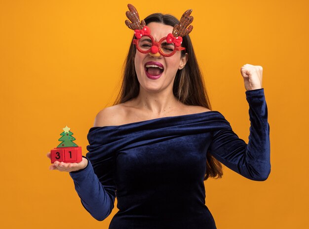 Excited with closed eyes young beautiful girl wearing blue dress and christmas glasses holding toy showing yes gesture isolated on orange wall