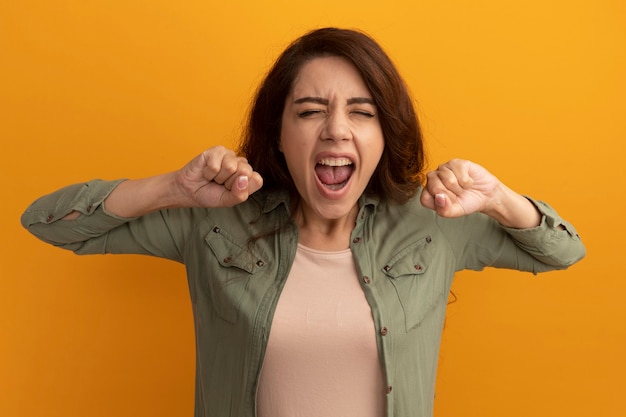 Excited with closed eyes young beautiful girl wearing olive green t-shirt holding fists isolated on yellow wall