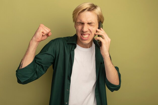 Excited with closed eyes young blonde guy wearing green t-shirt speaks on phone showing yes gesture