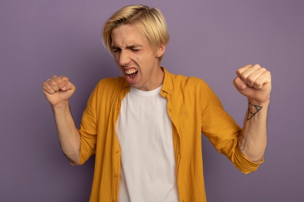 Free photo excited with closed eyes young blonde guy wearing yellow t-shirt showing yes gesture isolated on purple