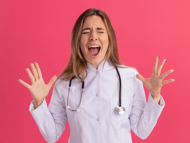 Excited with closed eyes young female doctor wearing medical robe with stethoscope spreading hands isolated on pink wall