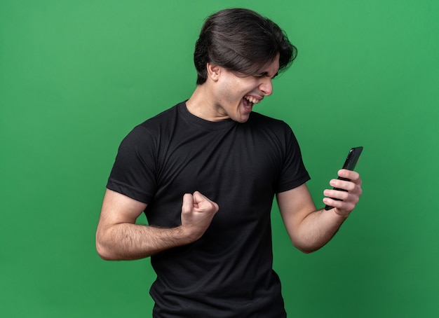 無料写真 目を閉じて興奮している若いハンサムな男は、緑の壁に分離されたはいジェスチャーを示す電話を保持している黒いtシャツを着ています