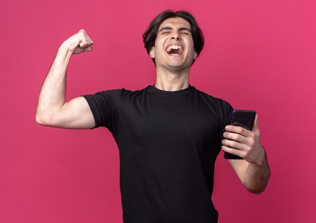 Excited with closed eyes young handsome guy wearing black t-shirt holding phone showing yes gesture isolated on pink wall