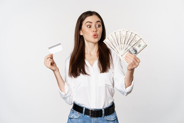 Excited woman holding credit card and money, looking amazed at cash, standing against white background