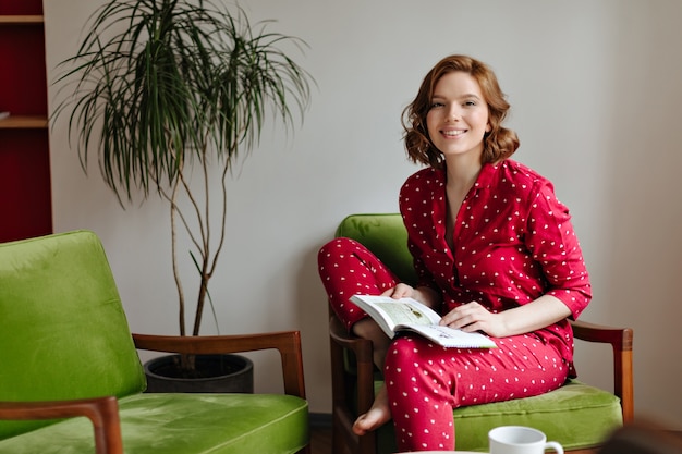 Excited woman in pajama sitting in armchair and looking at camera. Indoor shot of happy woman holding book.