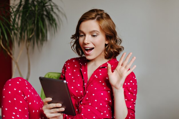 Excited woman in pajama waving hand during video call. Indoor shot of woman in sleepwear using digital tablet.