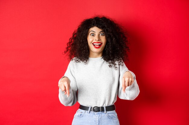 Excited woman pointing fingers down and smiling amazed, look here gesture, showing logo on red empty space.