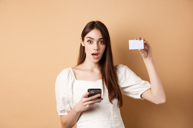 Возбужденная женщина показывает пластиковую кредитку и пользуется мобильным телефоном с отвисшей челюстью и задыхается от изумления ...