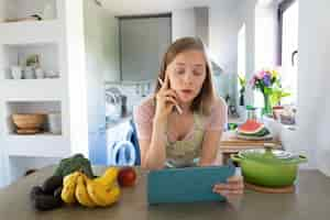 Free photo excited woman watching online cooking class in her kitchen, leaning on table, using tablet near saucepan and fresh fruits on counter. front view. cooking at home and healthy eating concept