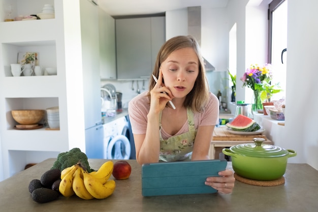 Взволнованная женщина смотрит онлайн-урок кулинарии на своей кухне, опираясь на стол, используя планшет возле кастрюли и свежие фрукты на прилавке. Передний план. Кулинария дома и концепция здорового питания