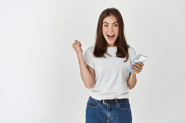 Excited woman winning prize on phone, rejoicing and looking at at front happy, scream of joy over white wall.