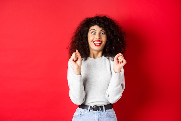 Excited woman winning prize, rejoicing and looking happy, smiling amazed, standing against red background