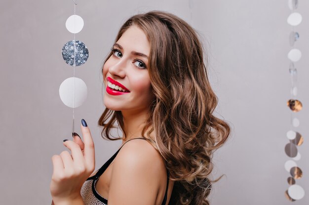 Excited woman with big light eyes posing with happy smile on dark wall. Indoor photo of joyful caucasian girl with brunette hair waiting for christmas party.