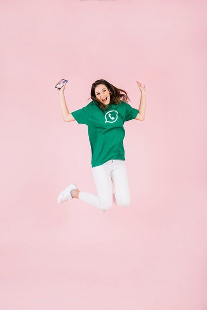 Excited woman with cellphone jumping over pink background