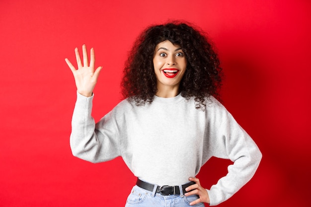 Возбужденная женщина с вьющимися волосами показывает номер пять пальцами, делая заказ, стоя на красном фоне