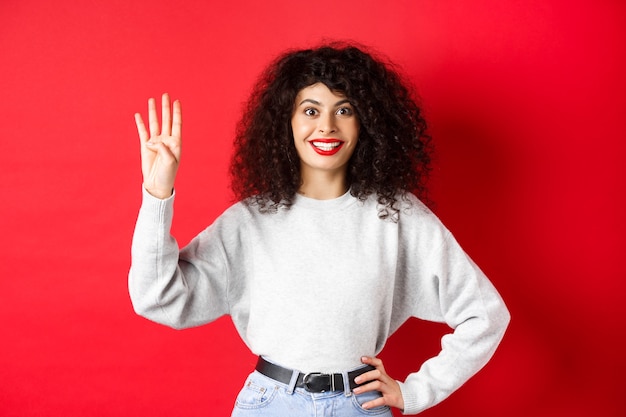 Donna eccitata con i capelli ricci che mostra il numero quattro con le dita, facendo ordine, in piedi su sfondo rosso