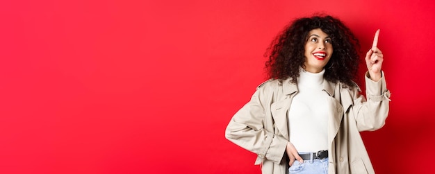 Excited woman with curly hair wearing trench coat pointing at upper right corner and smiling amazed