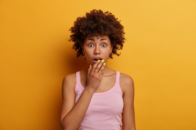 Excited wondered curly adult woman gasps astonishing, keeps hand on opened mouth, holds breath, hears something unbelievable, poses against yellow wall, reacts on latest unbelievable news