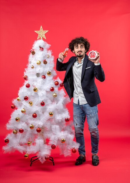 Excited young adult holding clock and raising a glass of wine and standing near Xsmas tree on red stock photo