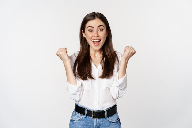 Excited young adult woman reacting to win, surprise news, screaming and cheering, triumphing, achieve goal and celebrating, standing over white background