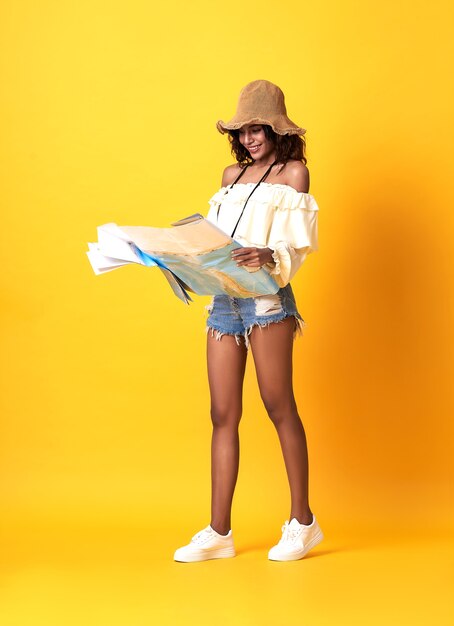 Excited young african woman dressed in summer clothes holding travel map guide over yellow wall.
