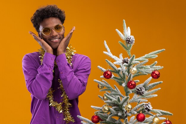 オレンジ色の壁で隔離された顔に手を保ちながら装飾されたクリスマスツリーの近くに立っている首の周りに見掛け倒しの花輪と眼鏡をかけている興奮した若いアフリカ系アメリカ人の男