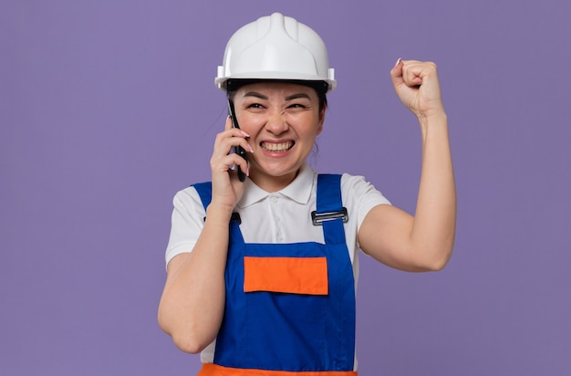 흰색 안전 헬멧을 쓴 흥분한 젊은 아시아 건축업자 소녀가 전화 통화를 하고 주먹을 들고 있다