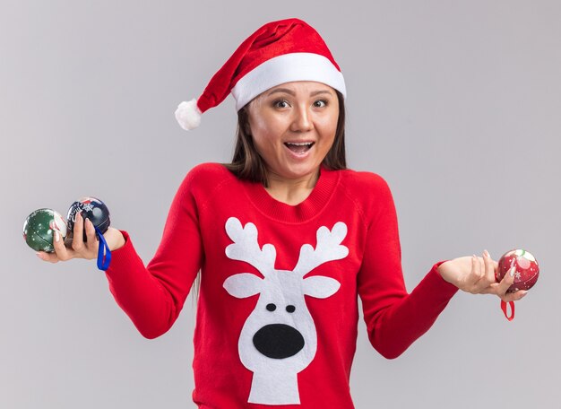 흰색 배경에 고립 된 크리스마스 트리 볼을 들고 스웨터와 크리스마스 모자를 쓰고 흥분된 젊은 아시아 여자