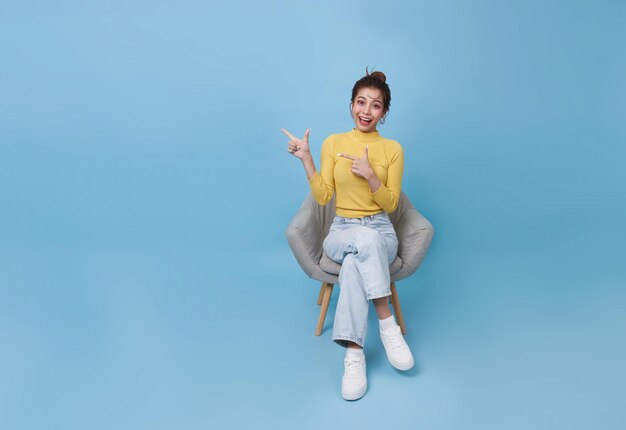 Excited young Asian woman sitting in armchair pointing fingers offering space