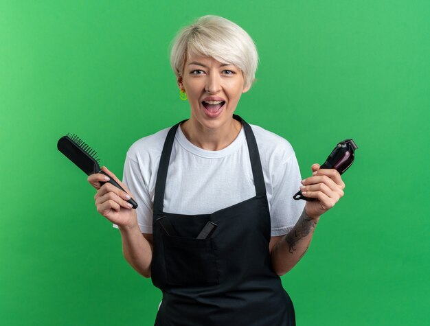 Excited young beautiful female barber in uniform holding hair clipper with comb isolated on green wall