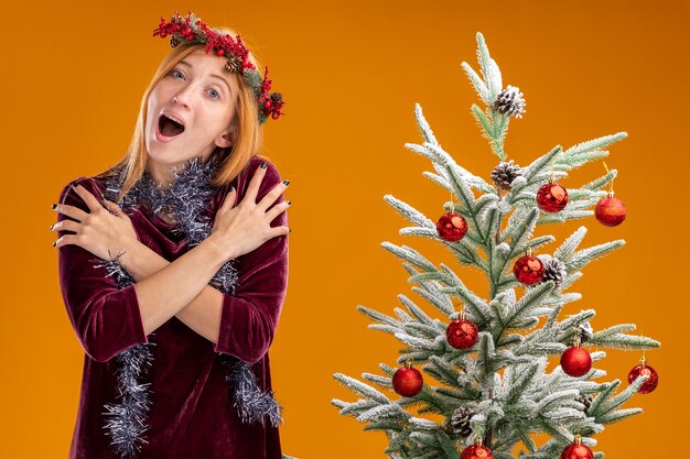 Excited young beautiful girl standing nearby christmas tree wearing red dress and wreath with garland on neck putting hands on shoulder isolated on orange wall