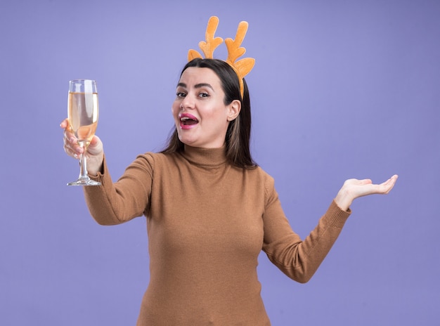 Возбужденная молодая красивая девушка в коричневом свитере с рождественским обручем для волос держит бокал шампанского, протягивая руку, изолированную на синей стене