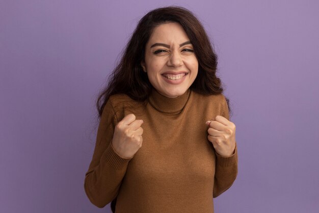 Excited young beautiful girl wearing brown turtleneck sweater holding fists isolated on purple wall