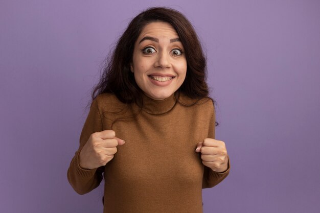 Excited young beautiful girl wearing brown turtleneck sweater showing yes gesture isolated on purple wall