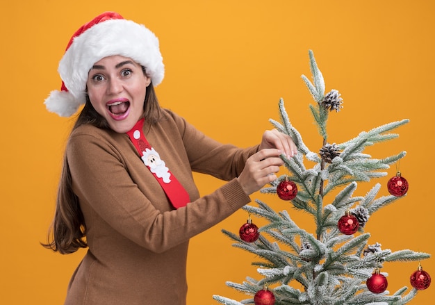 Excited young beautiful girl wearing christmas hat with tie standing nearby christmas tree isolated on orange background