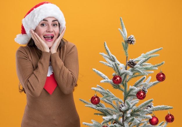 Excited young beautiful girl wearing christmas hat with tie standing nearby christmas tree putting hands on cheeks isolated on orange background