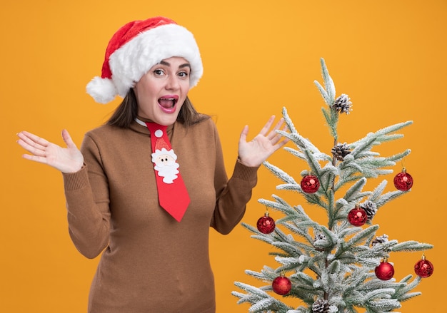 オレンジ色の背景で隔離の手を広げてクリスマスツリーの近くに立っているネクタイとクリスマス帽子をかぶって興奮した若い美しい少女
