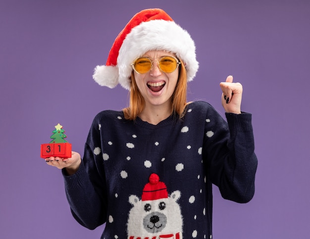 紫色の壁に分離されたはいジェスチャーを示すクリスマスのおもちゃを保持しているメガネとクリスマスセーターと帽子を身に着けている興奮した若い美しい少女