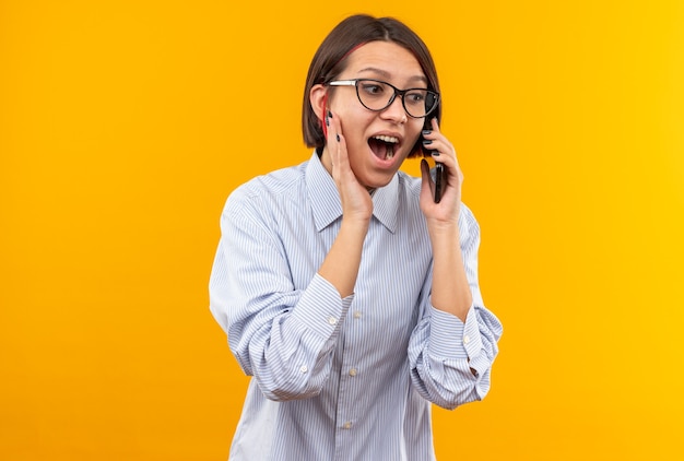Excited young beautiful girl wearing glasses speaks on phone putting hand on cheek isolated on orange wall
